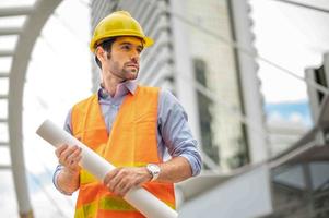 jong Kaukasisch Mens Holding een groot papier, vent vervelend licht blauw overhemd en jeans met oranje hesje en geel helm voor veiligheid in bouw Oppervlakte. foto