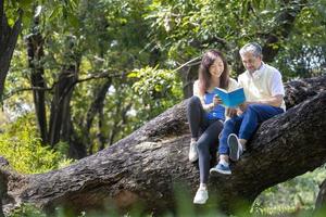 Aziatisch senior vader en dochter zittend en lezing een boek samen Aan de Afdeling van volwassen boom gedurende zomer voor fysiek en mentaal Gezondheid en levensduur concept foto