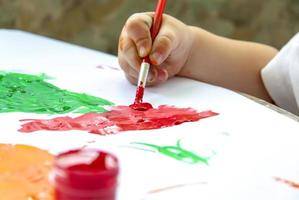 een kind hand- houdt een borstel en verven papier met groot slagen. uitstrijkjes van helder verf Aan wit papier. foto