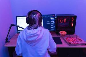 vrouw gamer of software ontwikkelaar zittend Bij de modern persoonlijk computer en aan het eten rommel voedsel Bij nacht in kamer met neon lichten foto