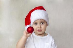 portret van een schattig weinig jongen in een wit t-shirt en de kerstman claus hoed. een jongen bewondert een rood Kerstmis bal. kinderen emoties. Kerstmis en nieuw jaar. foto