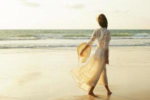 vrouw vervelend mooi wit jurk is wandelen Aan de strand gedurende zonsondergang foto