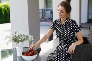 prachtig vrouw vervelend mooi jurk met een polka punt patroon zittend in een patio en aan het eten aardbei foto
