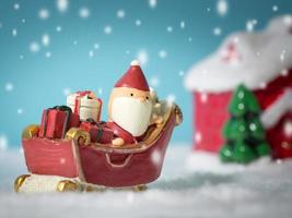 gelukkig de kerstman claus met cadeaus doos Aan de sneeuw slee gaan naar sneeuw huis. in de buurt sneeuw huis hebben sneeuwman en Kerstmis boom. de kerstman claus en sneeuw huis Aan de sneeuw de achtergrond is poeder blauw. foto