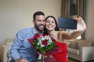 beeld van gelukkig jong paar nemen selfie foto met bloemen terwijl hebben een romantisch tijd Bij huis. lief paar vieren Valentijnsdag dag