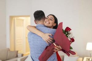 Valentijnsdag dag concept. gelukkig paar in liefde met. een jong liefhebbend paar vieren Valentijnsdag dag. geliefden knuffelen elk ander. knap Mens verrassingen zijn vriendin met rood rozen Bij huis foto