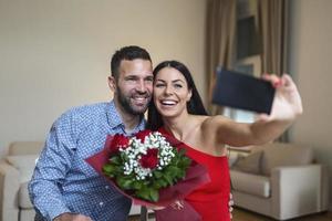 beeld van gelukkig jong paar nemen selfie foto met bloemen terwijl hebben een romantisch tijd Bij huis. lief paar vieren Valentijnsdag dag