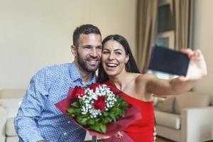 beeld van gelukkig jong paar nemen selfie foto met bloemen terwijl hebben een romantisch tijd Bij huis. lief paar vieren Valentijnsdag dag