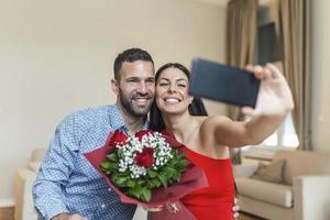 beeld van gelukkig jong paar nemen selfie foto met bloemen terwijl hebben een romantisch tijd Bij huis. lief paar vieren Valentijnsdag dag