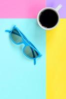 klein wit koffie kop en blauw zonnebril Aan structuur achtergrond van mode pastel blauw, geel, paars en roze kleuren papier in minimaal concept foto