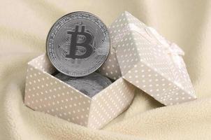 de zilver bitcoin leugens in een klein oranje geschenk doos met een klein boog Aan een deken gemaakt van zacht en pluizig licht oranje fleece kleding stof met een groot aantal van Verlichting vouwen foto