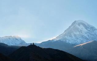 Georgië, monteren Kazbek in de stralen van de instelling zon, silhouet van de Gergeti drie-eenheid kerk over- dorp van stiefmoeder. historisch plaats van Georgië, toerist routes, berg klimmen, bedevaart foto