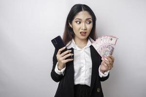 een verward jong zakenvrouw is vervelend een zwart pak, Holding haar telefoon en geld in Indonesisch roepia geïsoleerd door een wit achtergrond foto