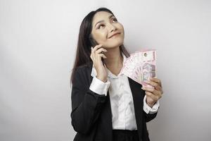 een gelukkig jong zakenvrouw is vervelend zwart pak, Holding haar telefoon en geld in Indonesisch roepia geïsoleerd door wit achtergrond foto