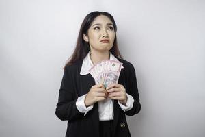 een verward jong zakenvrouw is vervelend zwart pak en Holding contant geld geld in Indonesisch roepia geïsoleerd door wit achtergrond foto