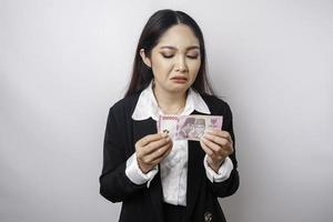 een portret van een Aziatisch zakenvrouw vervelend een zwart pak en Holding geld in Indonesisch roepia geïsoleerd door wit achtergrond looks depressief foto