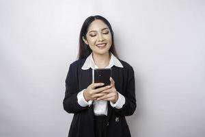 een portret van een gelukkig Aziatisch zakenvrouw is glimlachen en Holding haar smartphone vervelend een zwart pak geïsoleerd door een wit achtergrond foto