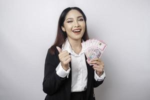 opgewonden Aziatisch zakenvrouw vervelend zwart pak geeft duimen omhoog hand- gebaar van goedkeuring en Holding geld in Indonesisch roepia, geïsoleerd door wit achtergrond foto