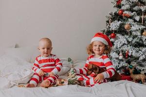 kinderen in rood en wit pyjama zittend in bed delen Kerstmis snoepgoed met elk andere en hun hond. broer en zus, jongen en meisje vieren de nieuw jaar. ruimte voor tekst. hoog kwaliteit foto