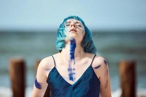 artistieke blauwharige vrouw performancekunstenaar besmeurd met blauwe gouacheverf dansend op het strand foto