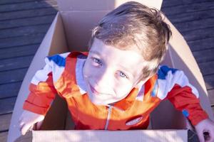 gelukkig jongen met blond haar- klimt uit van een groot karton doos, glimlacht, geschenk, verrassing foto