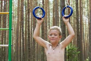 portret van een schattig jongen trekken zichzelf omhoog Aan gymnastiek- ringen in natuur tegen de achtergrond van een Woud foto
