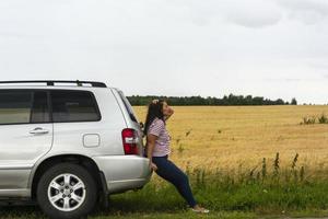 een gebroken auto. een jong vrouw staat Aan de weg door een gebroken auto tegen de achtergrond van een geel veld. foto