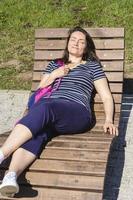 jong brunette aan het liegen Aan een houten sjees longue in een stad park Aan een zonnig dag, stad vakantie, jong aantrekkelijk vrouw genieten in de zon foto