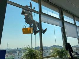 een mannetje venster wasmachine arbeider, industrieel klimmer blijft hangen Aan een hoog gebouw, wolkenkrabber en wast groot glas ramen voor netheid hoog bovenstaand een groot stad in een kantoor bedrijf gebouw foto