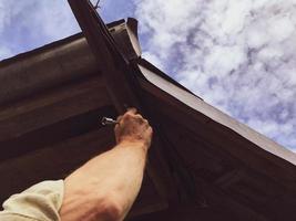 een Mens reparaties de dak met metaal gereedschap Bij een hoogte. fabricage van een dak in een houten huis. vulling gaten in een woon- gebouw, bescherming van neerslag. draaien de bouten, blauw lucht foto