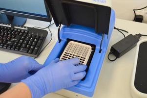 een dokter in een blauw steriel handschoen zet een bord met een reagens in de inrichting naar uitvoeren de studie. laboratorium analysator. veneus bloed test foto