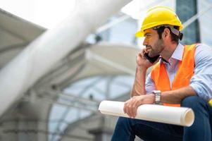 Kaukasisch Mens ingenieur gebruik een smartphone voor praten, vervelend oranje hesje en groot moeilijk hoed, en de andere hand- Holding de wit verdieping plan in de plaats werk van de centrum stad. foto