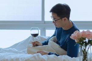 een persoon lezing boek Aan bed met een glas van rood wijn. digitaal ontgiften en sociaal verbinding verbreken concept. foto