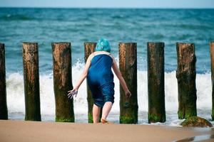 blauwharige vrouw performance artiest in blauwe jurk staande op het strand met kwast, achteraanzicht foto