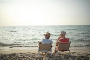 twee ouderen mannen en Dames zitten stoel Bij de strand pratend en aan het kijken de zon en de zee Aan hun zomer vakantie en ze glimlach en genieten hun vakantie. foto