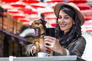 jong vrouw met haar hond in huisdier vriendelijk cafetaria. meisje drinken koffie Bij lokaal koffie winkel met haar huisdier hond nemen een selfie met mobiel telefoon foto