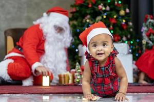 afro-amerikaanse baby heeft plezier met spelen met speelgoed terwijl de kerstman een cadeau voorbereidt en achter de kerstboom zit voor het concept van de seizoensviering foto
