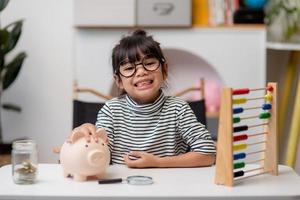 klein Aziatisch meisje dat geld spaart in een spaarvarken, leert over sparen, kind spaart geld voor toekomstig onderwijs. geld, financiën, verzekeringen en mensenconcept foto