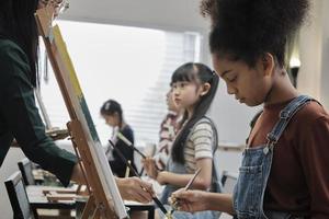 Afrikaanse Amerikaans meisje concentraten Aan acryl kleur afbeelding schilderij Aan canvas met studenten groep in kunst klas, creatief aan het leren met talenten en vaardigheden in elementair school- studio onderwijs. foto