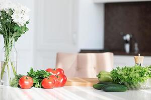 ingrediënten en gereedschap voor vers gezond groenten salade. de concept van huis Koken. groen salade, komkommers, rood tomaten, bloemen Aan de tafel Aan wit keuken. zonnig keuken. kopiëren ruimte, achtergrond foto