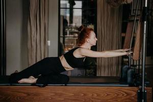 roodharige vrouw doet stretching-routine op cadillac-machine of trapeze-tafel foto