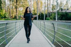 sportieve Europese vrouw in zwart t-shirt en legging, loopt over de brug, houdt twee halters vast, werkt aan het trainen van biceps, traint buiten, heeft een doordachte uitdrukking, poseert met sportuitrusting foto