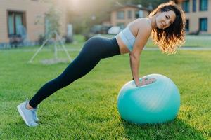 horizontaal schot van mooie slanke vrouw doet fysieke oefeningen met fitnessbal heeft een perfect figuur draagt sportkleding poses op groen gazon buiten geniet van fysieke training. pilates en yogaconcept foto