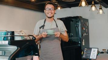 portret jong Aziatisch vent barista serveerster Holding koffie kop gevoel gelukkig Bij stedelijk cafe. Azië klein bedrijf eigenaar jongen in schort kom tot rust toothy glimlach op zoek naar camera staan Bij teller in koffie winkel. foto