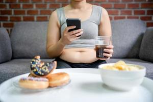 te veel eten en gulzigheid concept. zwaarlijvig vrouw zittend Aan sofa aan het eten ongezond voedsel foto