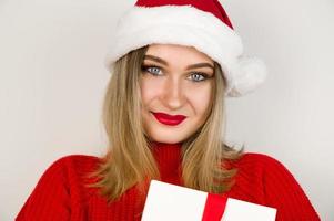 detailopname portret van vrouw blond in rood trui met de kerstman hoed glimlachen en Holding Geschenk. kerst vooravond, feestdagen concept foto