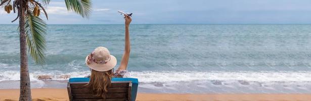 zomer strand vakantie vakantie reis concept, gelukkig jong Aziatisch vrouw met hoed ontspannende Aan strand stoel en Holding vliegtuig model- vliegend over- blauw lucht. foto