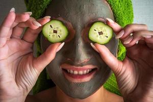 mooi jong vrouw met gelaats masker Aan haar gezicht Holding plakjes van komkommer. foto