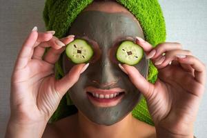 mooi jong vrouw met gelaats masker Aan haar gezicht Holding plakjes van komkommer. foto