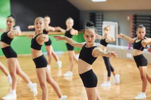 staand en aan het doen gesynchroniseerd beweegt. groep van vrouw kinderen beoefenen atletisch opdrachten samen binnenshuis foto